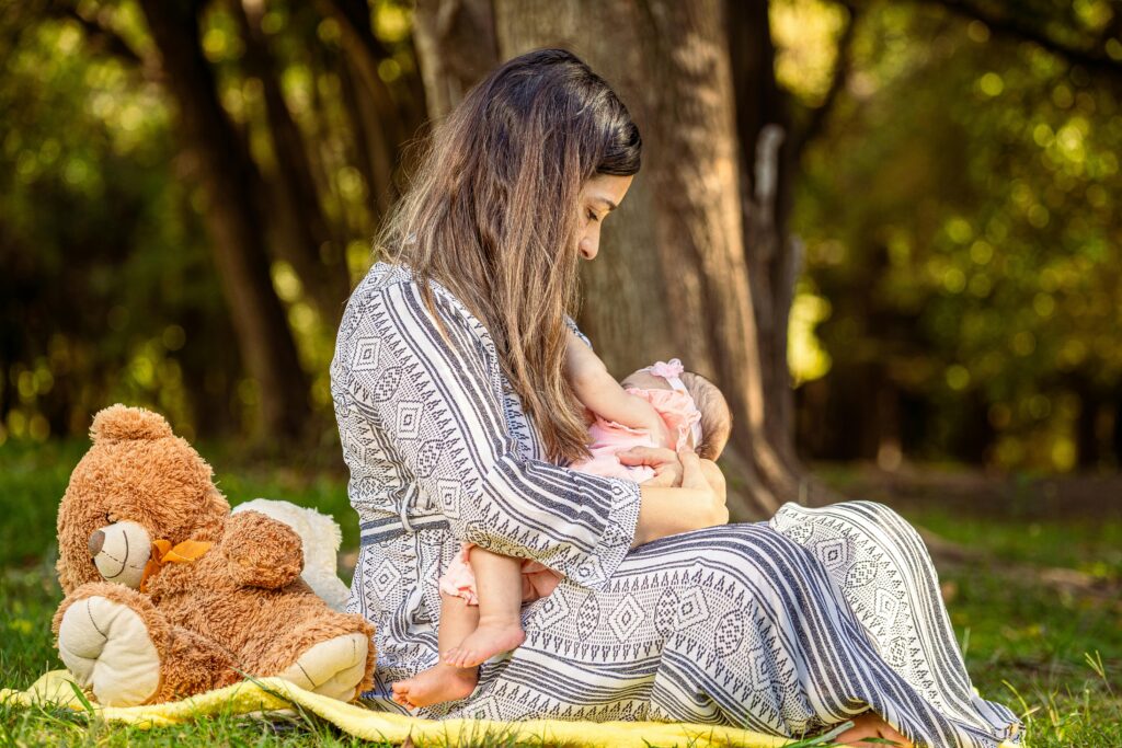 Tender moment of a mother breastfeeding her baby outdoors with a teddy bear nearby.