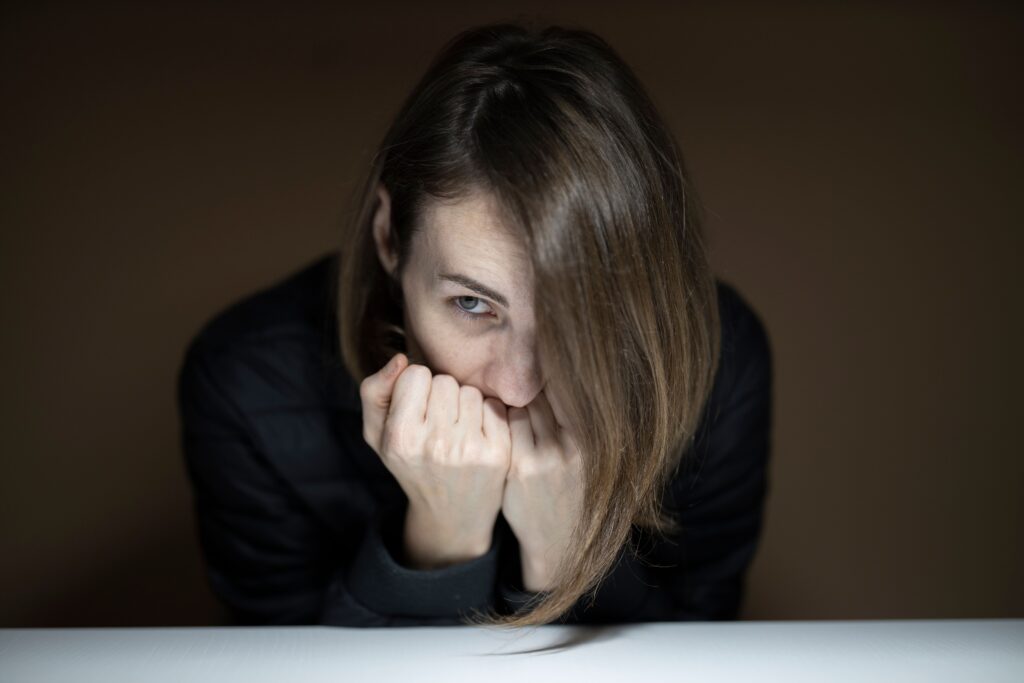 A young woman sits indoors, partially obscuring her face, evoking a thoughtful and introspective mood.